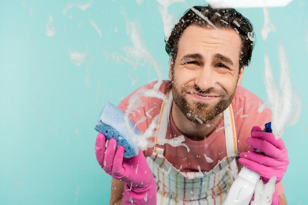 Upset man in rubber gloves holding detergent and sponge near glass isolated on blue  - Photo, Image