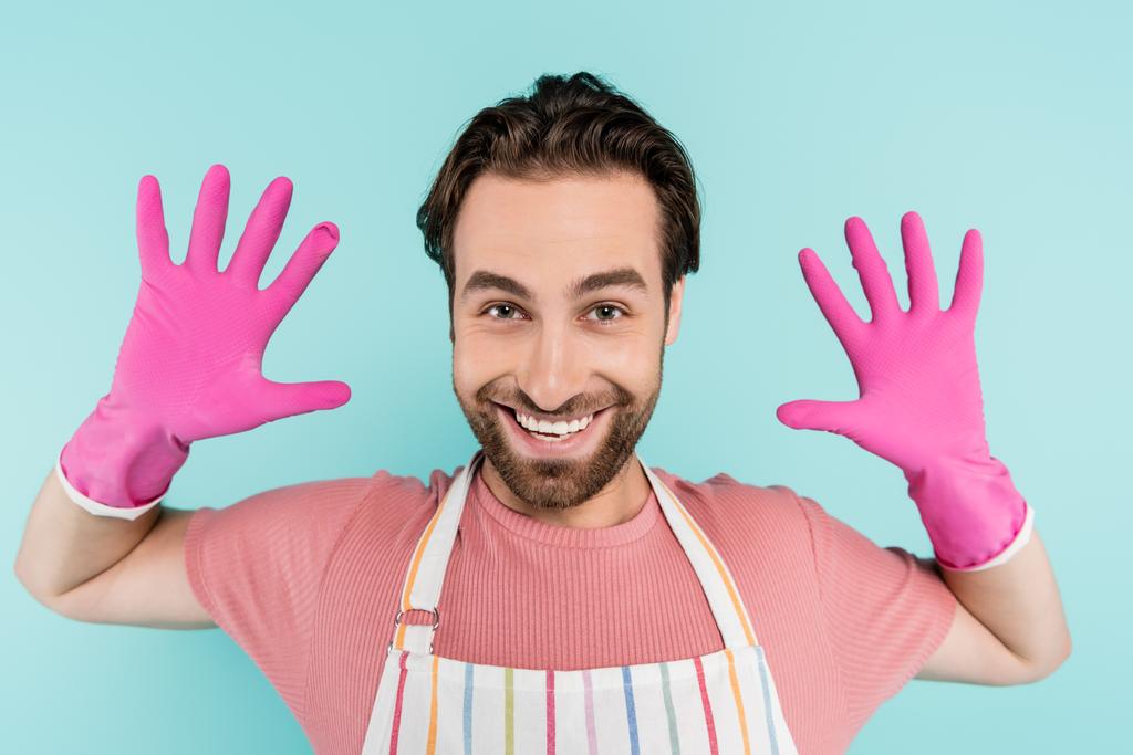 Positive man in rubber gloves and apron looking at camera isolated on blue  - Photo, Image