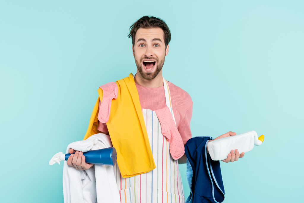 Aufgeregter Mann in Schürze hält Waschmittel und Kleidung isoliert auf blauem Grund  - Foto, Bild