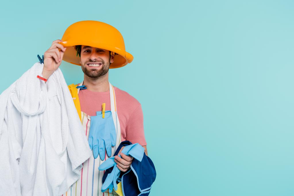 Fröhlicher Mann in Wäscheklammern mit Waschschüssel auf dem Kopf und isolierter Kleidung auf blauem Grund  - Foto, Bild