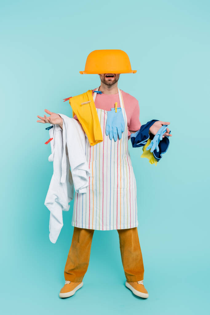 Shocked unmarried man in clothespins, holding clothes and wash bowl on head on blue background  - Photo, Image