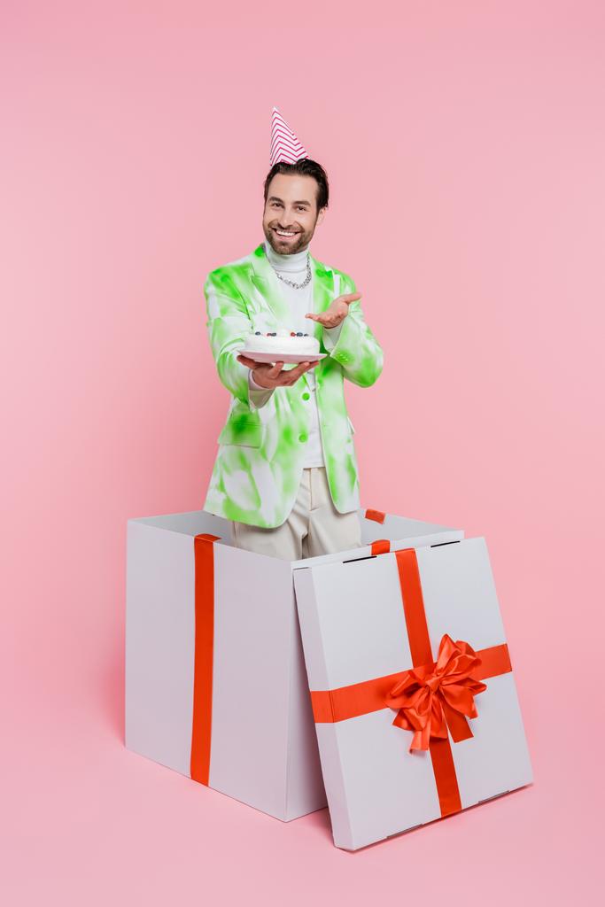 Pozitivní muž ve straně čepice ukazující na narozeninový dort v dárkové krabici na růžovém pozadí  - Fotografie, Obrázek