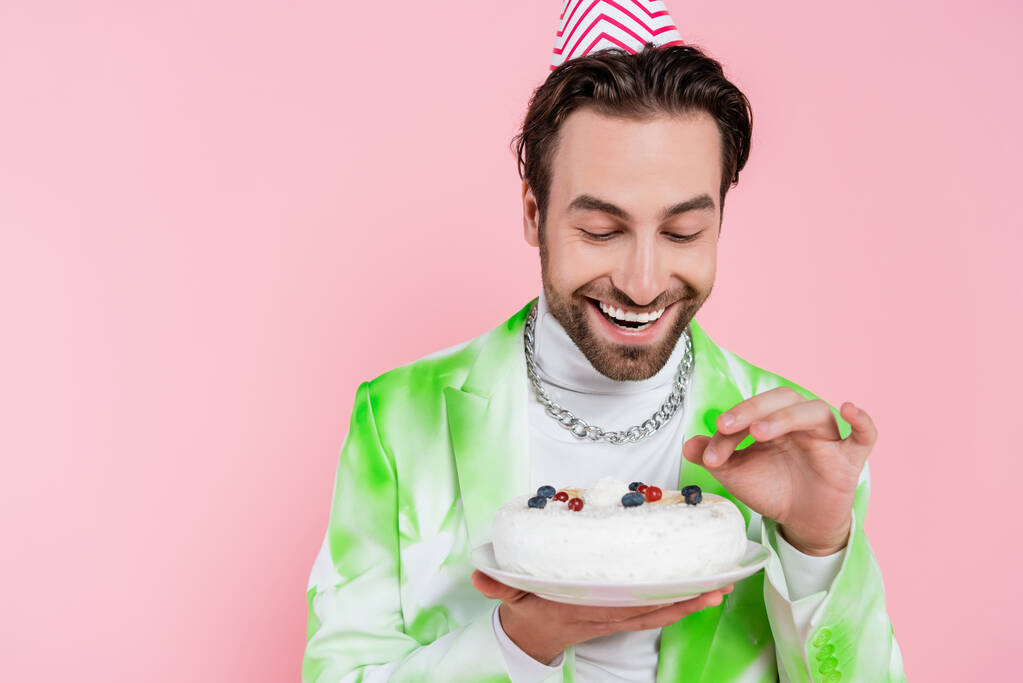Hombre positivo en gorra de fiesta sosteniendo pastel de cumpleaños aislado en rosa  - Foto, Imagen