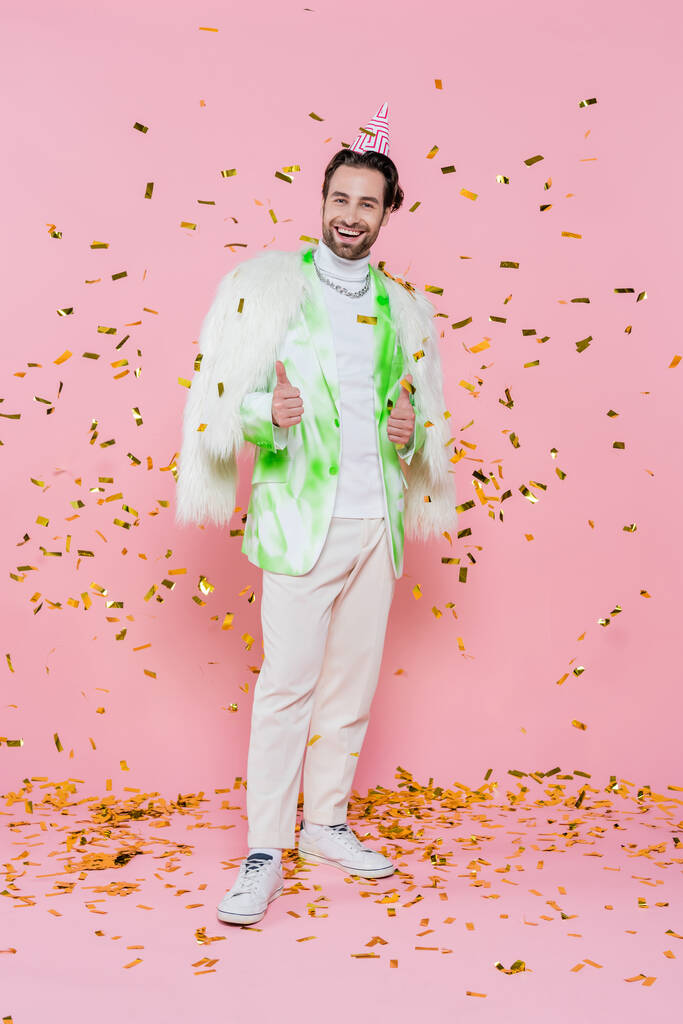 Vrolijke man in harige jas en feestmuts tonen als onder confetti op roze achtergrond  - Foto, afbeelding