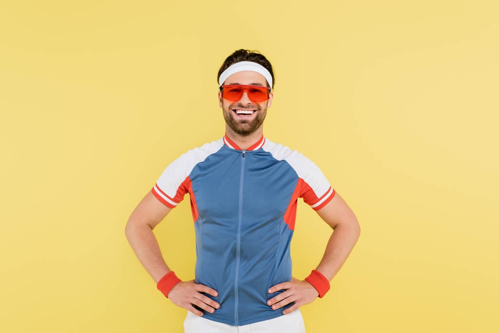 Veselý sportovec ve slunečních brýlích drží ruce na bocích izolované na žluté  - Fotografie, Obrázek