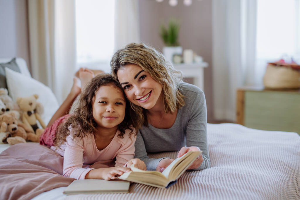 Glückliche Mutter mit ihrer kleinen Tochter, die zu Hause auf dem Bett liegt und Buch liest. - Foto, Bild
