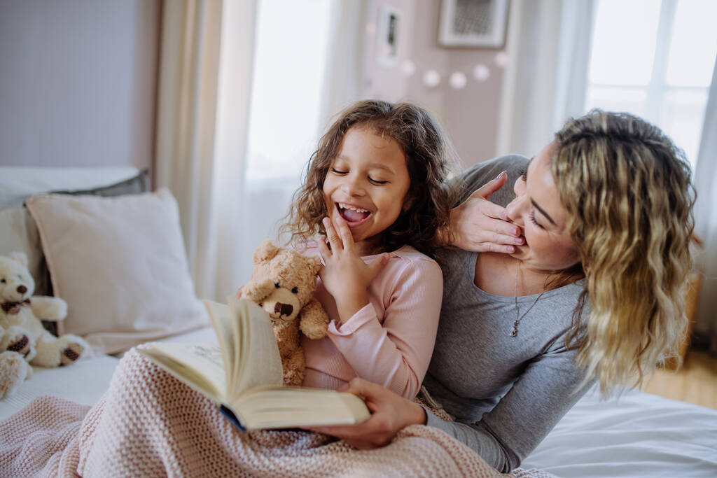 Счастливая мать со своей маленькой дочерью сидит на кровати и читает книгу дома. - Фото, изображение