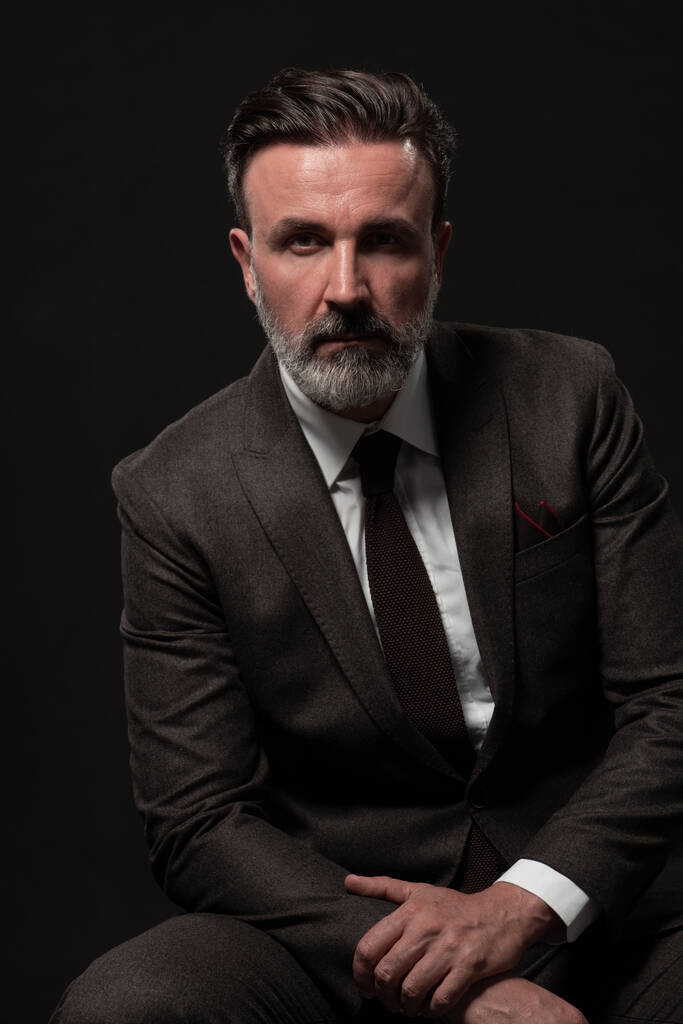 Portrét stylového elegantního staršího podnikatele s vousy a příležitostným obchodním oblečením ve fotoateliéru izolovaném na tmavém pozadí gestikulující rukama - Fotografie, Obrázek