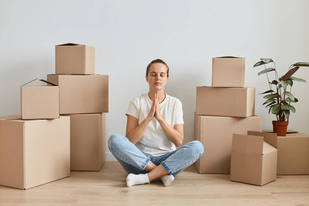 Calm attractive woman wearing white t shirt sitting on floor in lotus pose surrounded with cardboard boxes with belongings, practicing yoga to relax. - Photo, Image