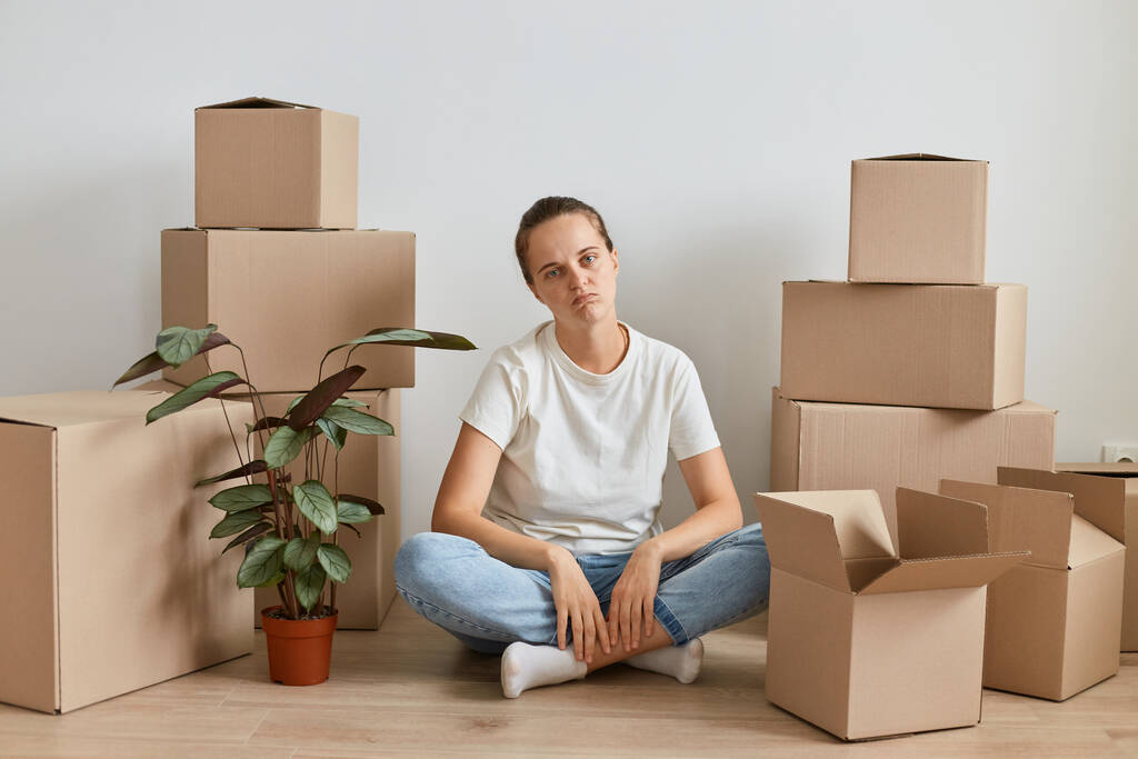 Image of bored tired woman wearing white t shirt sitting on floor surrounded with cardboard boxes with belongings, being exhausted during moving to a new apartment. - Photo, Image