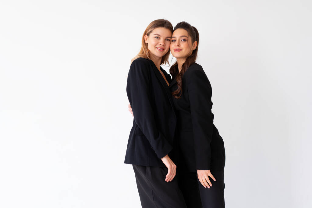 Концепция партнерства в бизнесе. Две молодые бизнесвумен одеты черный официальный костюм стоя студии изолированный белый фон. Белая деловая женщина, щека к щеке, улыбка - Фото, изображение