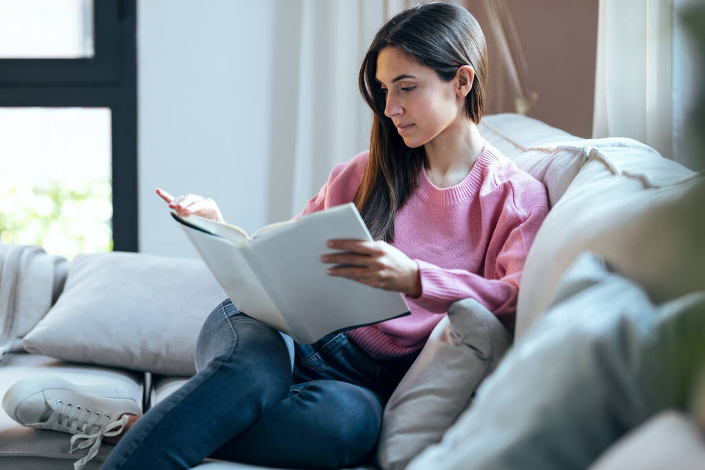 Foto de una mujer joven y bonita leyendo un libro mientras está sentada en el sofá en casa
. - Foto, Imagen