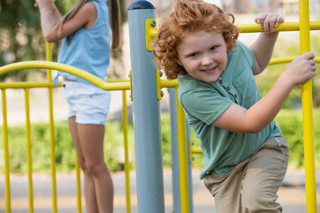 excited boy looking at camera while playing near blurred sister on playground - Photo, Image