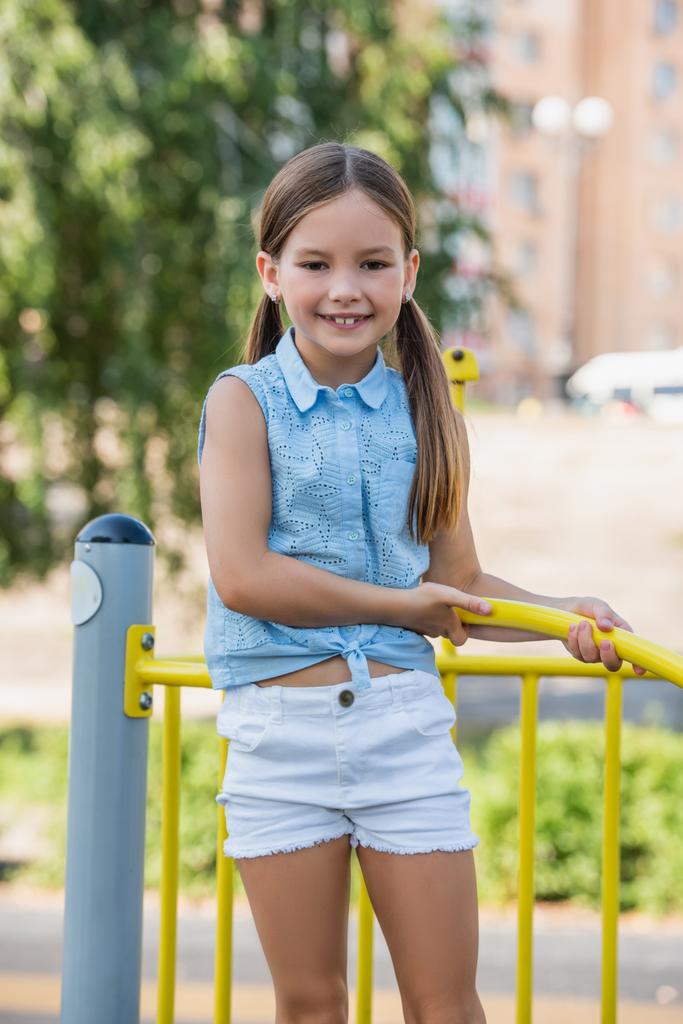 menina alegre em blusa sem mangas e curta sorrindo para a câmera no parque infantil - Foto, Imagem