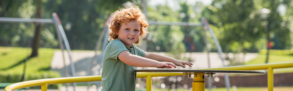roodharige jongen op zoek naar camera tijdens het rijden carrousel in de zomer park, banner - Foto, afbeelding
