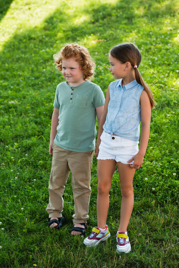 full length view of brother and sister in summer clothes standing on green lawn - Photo, Image