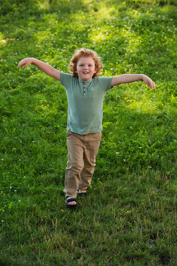 visão comprimento total do menino feliz com as mãos estendidas andando no gramado verde - Foto, Imagem