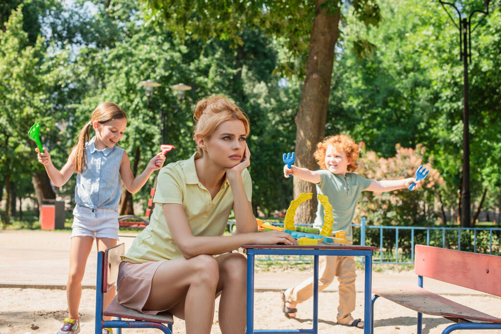 traurige Frau, die neben fröhlichen Kindern sitzt und wegsieht, die mit Spielzeugharken spielen - Foto, Bild