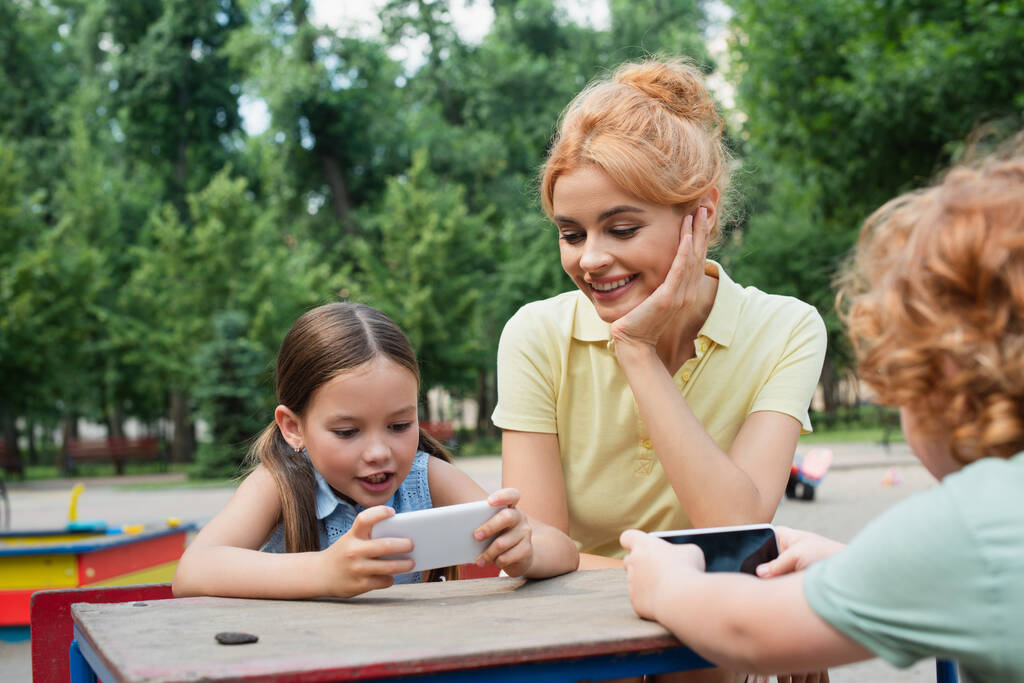 Glückliche Frau lächelt in der Nähe von Kindern mit Smartphones im Park - Foto, Bild