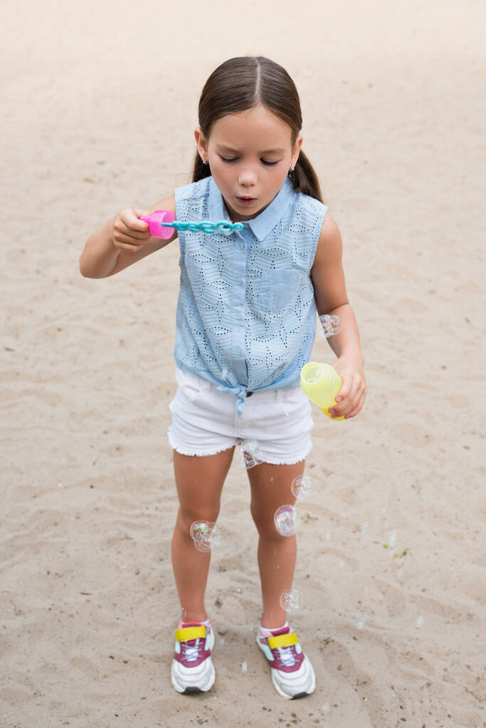 Вид в полный рост девушки в шортах, надувающей мыльные пузыри на улице - Фото, изображение
