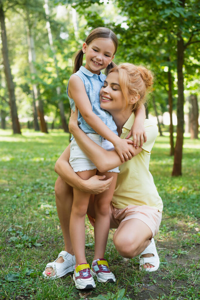 vista completa de la madre y la hija feliz en ropa de verano abrazando en el parque - Foto, Imagen