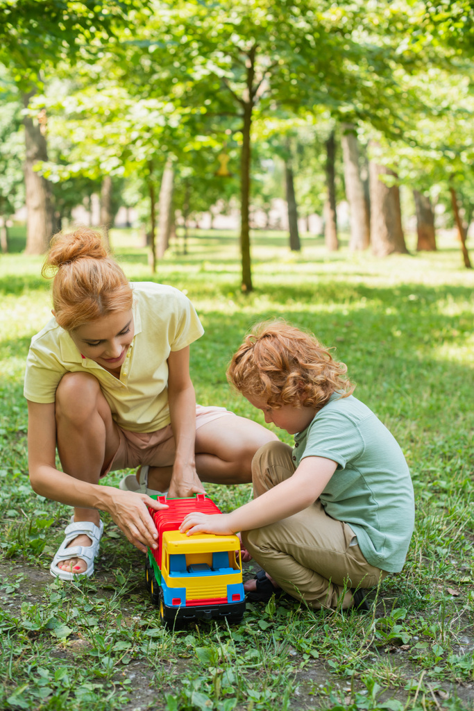 夏の公園の緑の芝生でおもちゃのトラックで遊んでいる赤毛の母親と息子 - 写真・画像