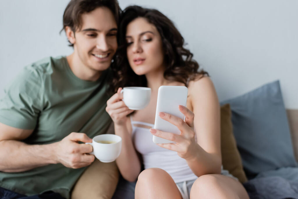 blurred couple with cups looking at smartphone in bedroom - Photo, Image