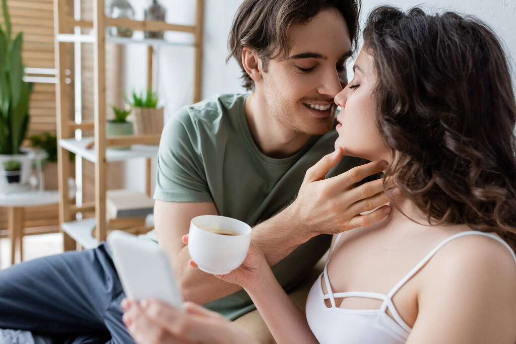 happy man kissing curly girlfriend with cup of coffee and smartphone - Photo, Image
