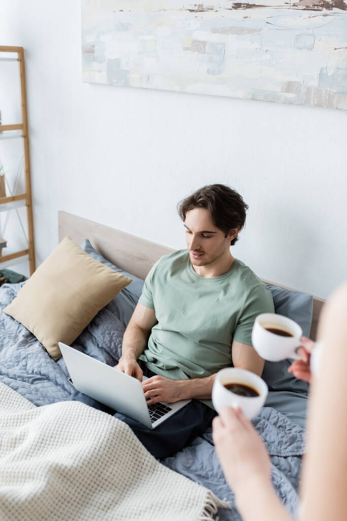 vista de ángulo alto de la mujer sosteniendo tazas de café cerca de novio independiente utilizando el ordenador portátil en la cama  - Foto, imagen