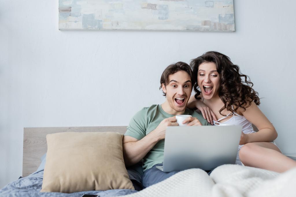 Yatak odasında dizüstü bilgisayara bakan heyecanlı genç çift. - Fotoğraf, Görsel