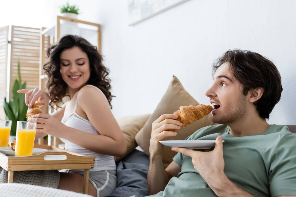 man eating croissant near happy woman during breakfast in bed - Photo, Image