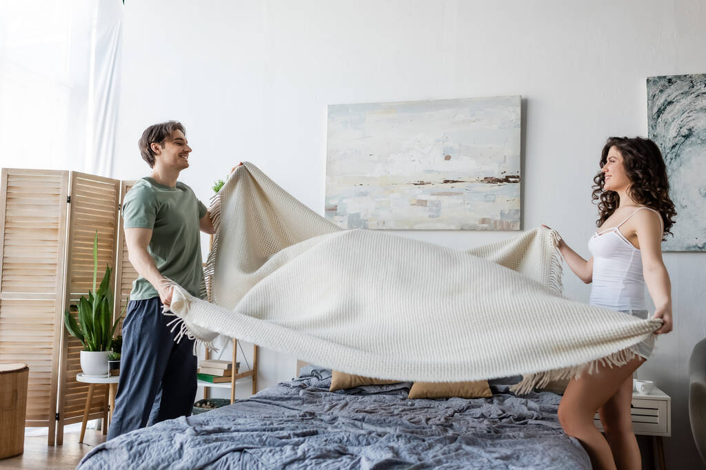 πλαϊνή άποψη του χαμογελαστού νεαρό ζευγάρι που καλύπτει κρεβάτι με κουβέρτα  - Φωτογραφία, εικόνα