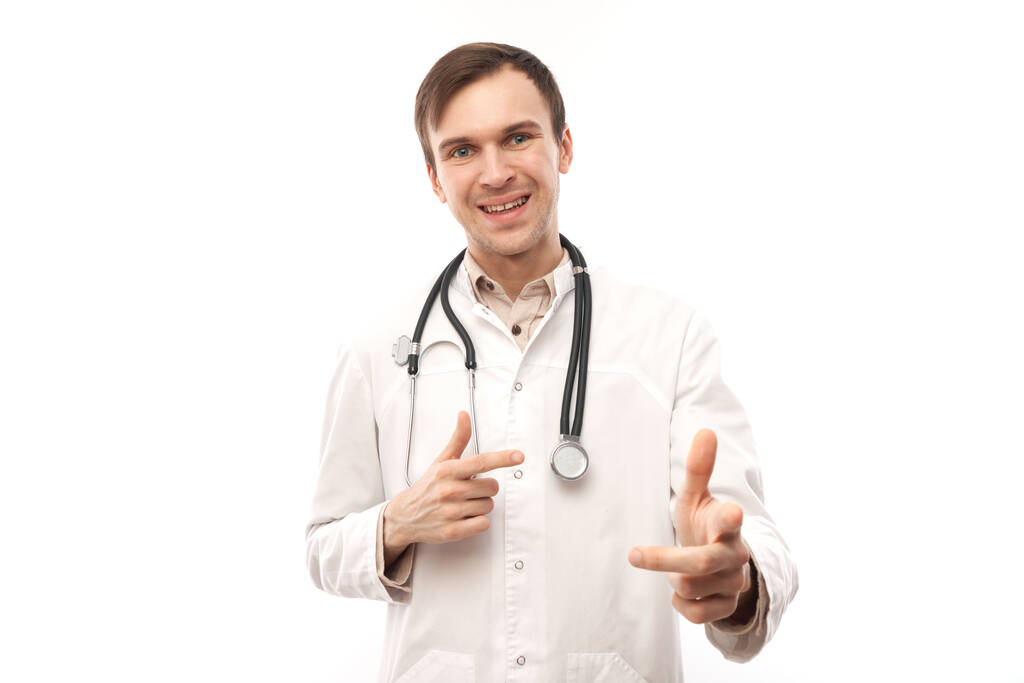 Porträt eines freundlichen, positiven jungen Arztes in weißem Arztkittel wählt dich aus, zeigt mit dem Finger auf die Kamera isoliert auf weißem Hintergrund - Foto, Bild