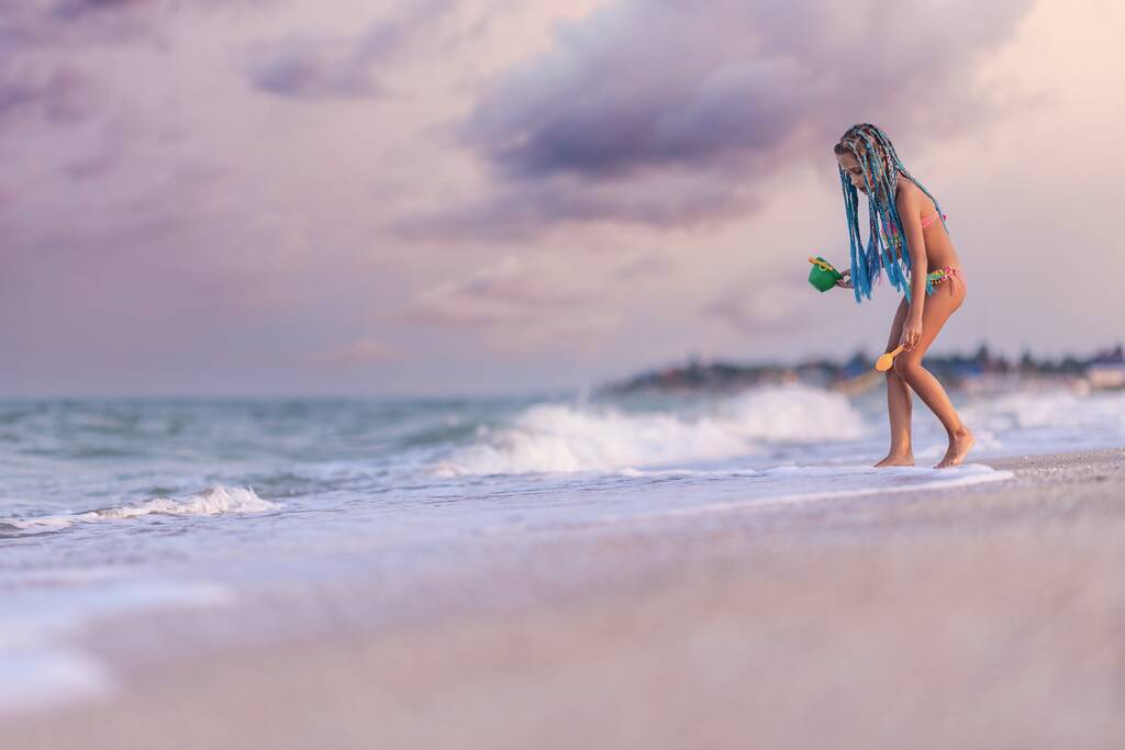 Ein Mädchen mit afrikanischen Zöpfen im Sommerkostüm spielt am Strand mit Muscheln am Meer mit Wellen unter sonnigem Sonnenuntergang - Foto, Bild