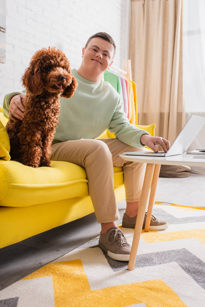 Smiling teenager with down syndrome petting poodle and using laptop on couch  - 写真・画像