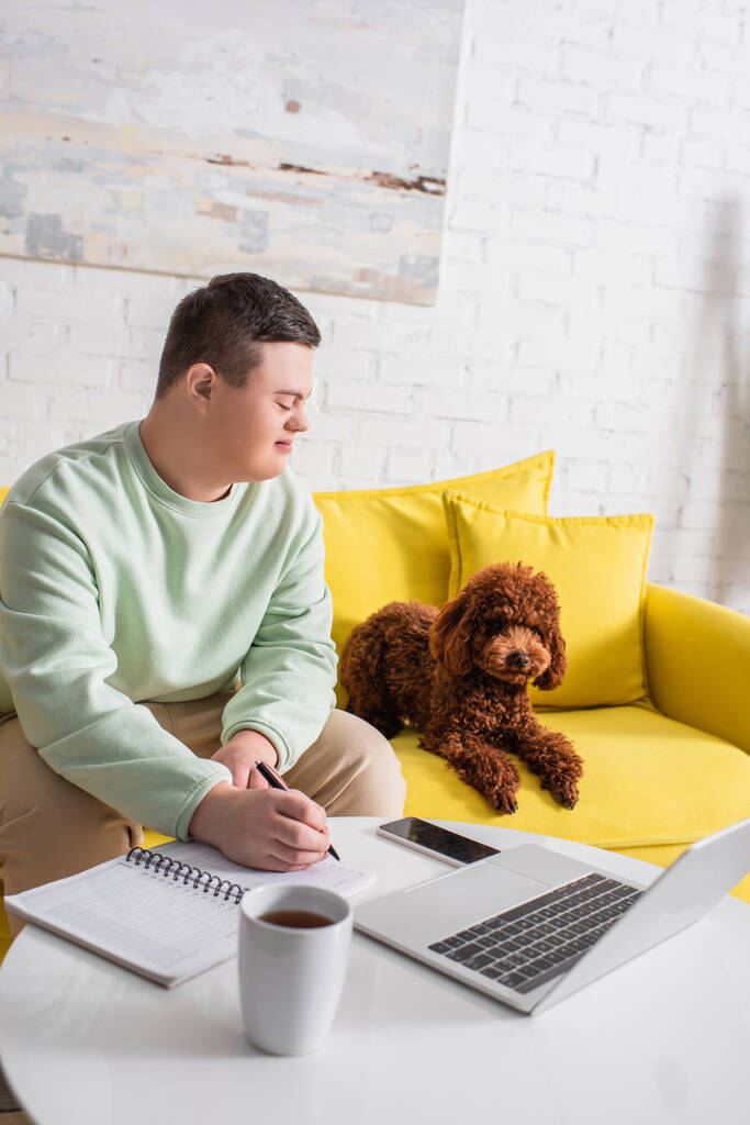 Side view of smiling teenager with down syndrome writing on notebook and looking at poodle near gadgets at home  - Photo, Image