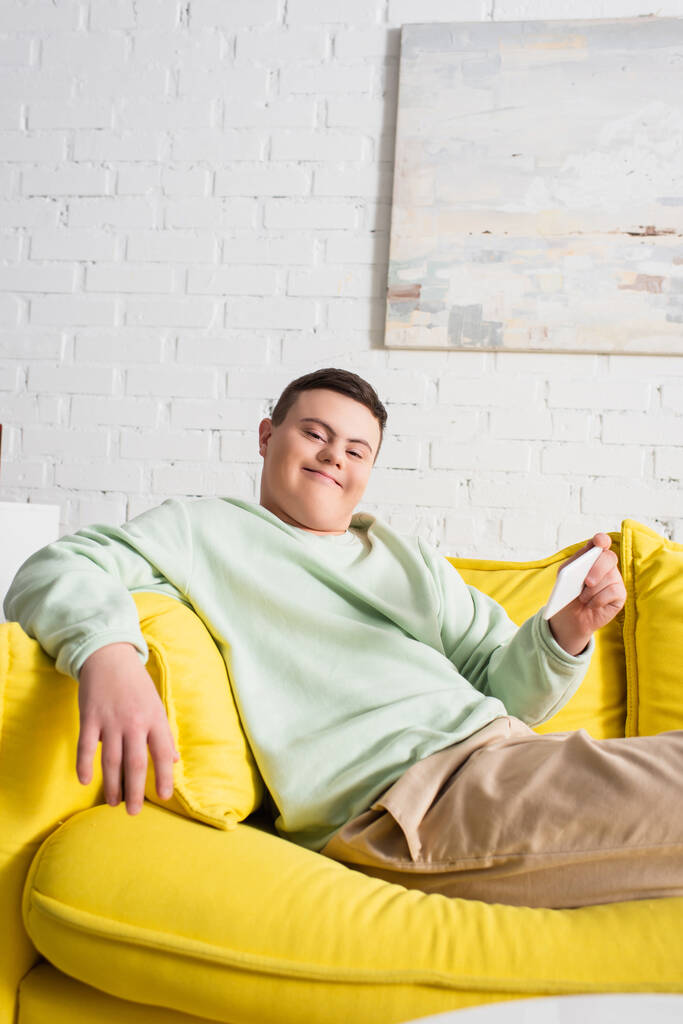 Down sendromlu pozitif bir genç evde kanepede otururken elinde akıllı telefon tutuyor.  - Fotoğraf, Görsel