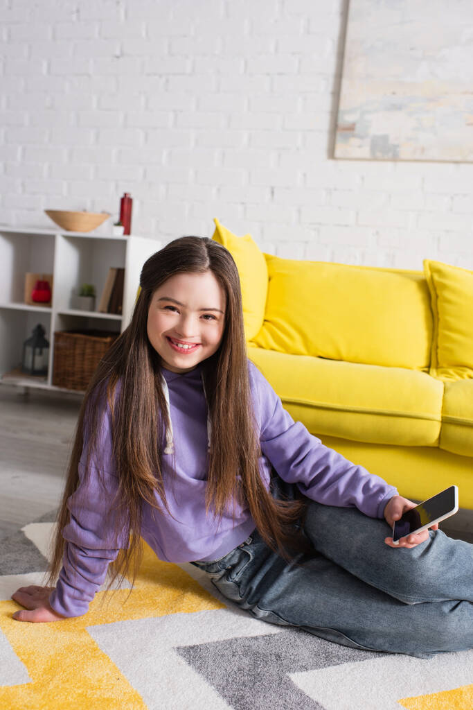 Menina alegre com síndrome de down segurando celular enquanto sentado no chão em casa  - Foto, Imagem