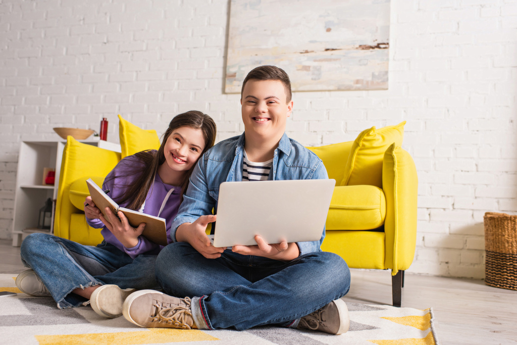Позитивные подростки с синдромом Дауна держат ноутбук и ноутбук на полу дома  - Фото, изображение