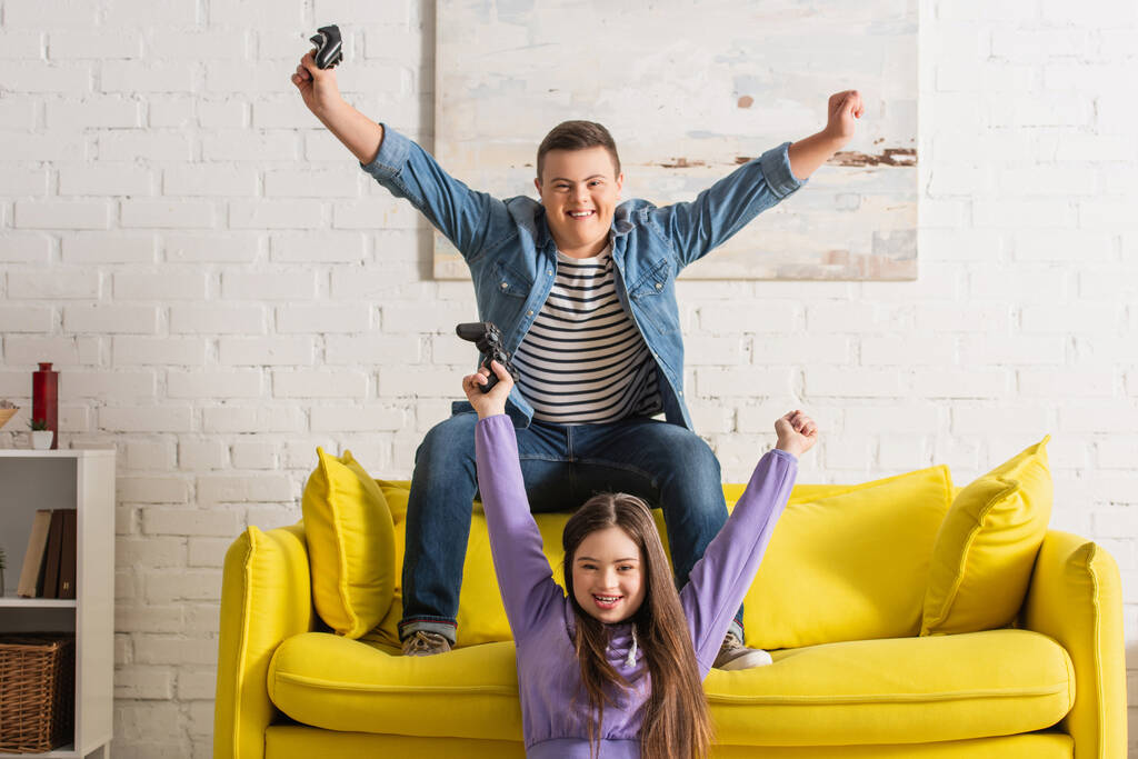 KYIV, UKRAJINA - 21. ČERVENCE 2022: Veselí teenageři s Downovým syndromem ukazujícím ano gesto při hraní videohry doma  - Fotografie, Obrázek