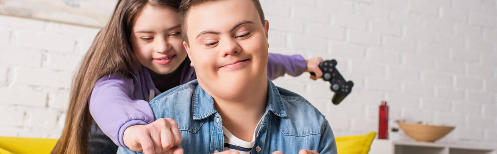 KYIV, UKRAJINA - 21. ČERVENCE 2022: Teen girl with down syndrome holding rozmazané joystick near friend at home, banner  - Fotografie, Obrázek