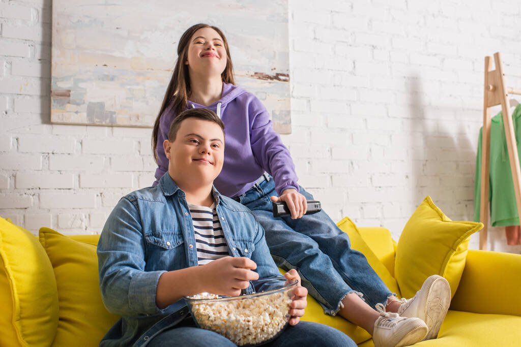Smiling teenager with down syndrome holding popcorn near friend with remote controller at home  - 写真・画像