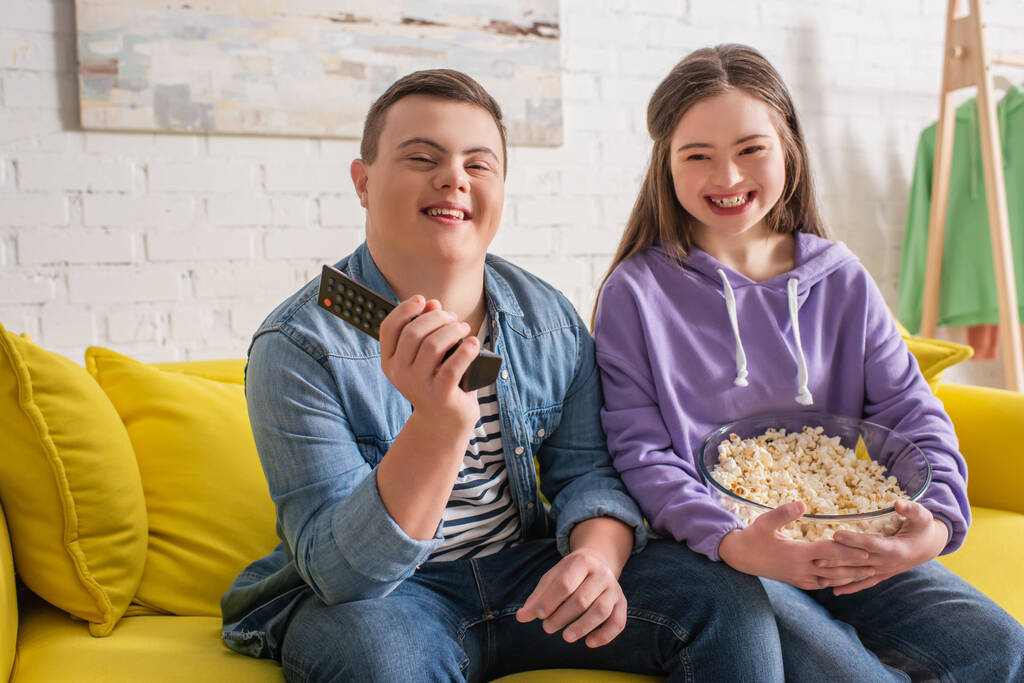 Happy teenagers with down syndrome holding remote controller and popcorn on couch  - 写真・画像