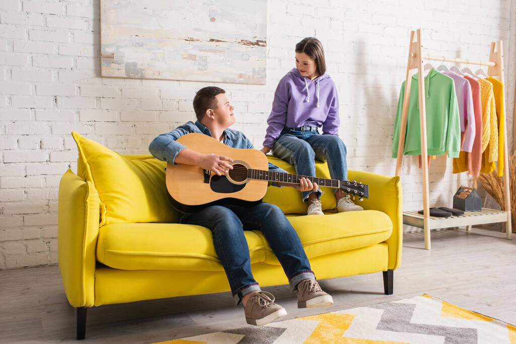 Adolescente tocando la guitarra acústica y mirando a un amigo con síndrome de Down en casa  - Foto, Imagen