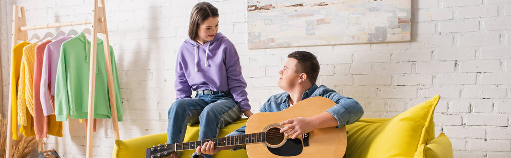 tiener meisje met down syndroom zitten in de buurt vriend spelen akoestische gitaar thuis, banner  - Foto, afbeelding