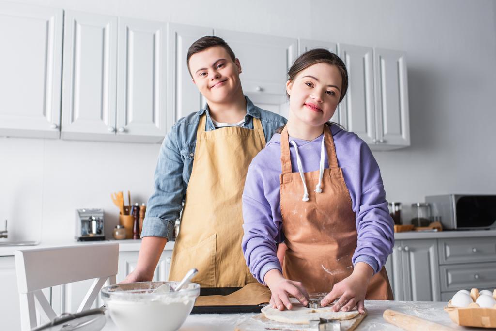 Alegre pareja con síndrome de Down mirando a la cámara mientras cocina en la cocina  - Foto, imagen