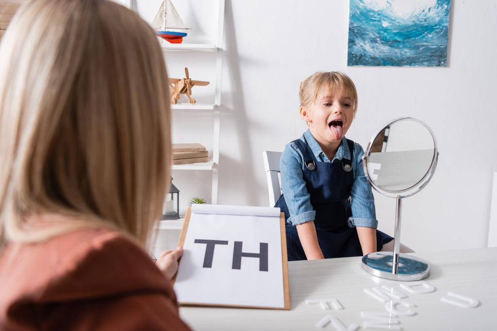 Pupil sticking out tongue near mirror and blurred speech therapist in classroom  - Фото, изображение