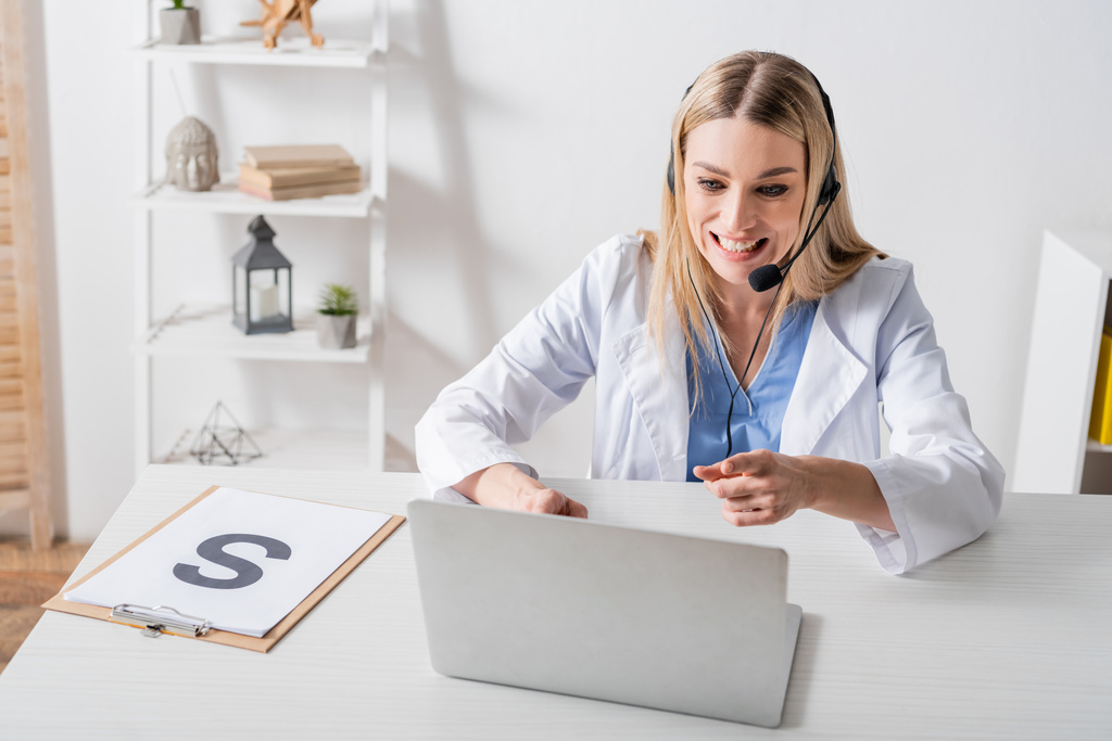 Smiling speech therapist in headset pointing at laptop during video call near clipboard in consulting room   - Photo, Image
