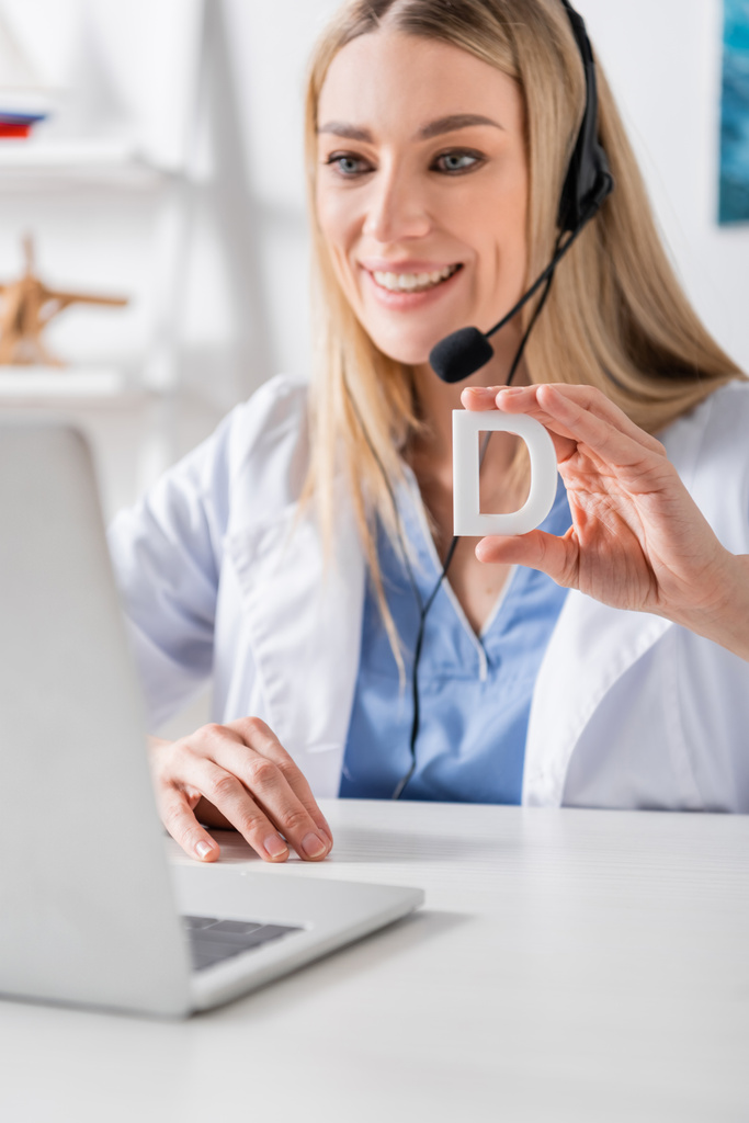 Smiling logopedist in headset holding letter near laptop during video call in consulting room  - Photo, Image