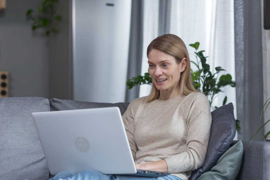 Женщина дома сидит на диване, улыбается и работает на ноутбуке - Фото, изображение